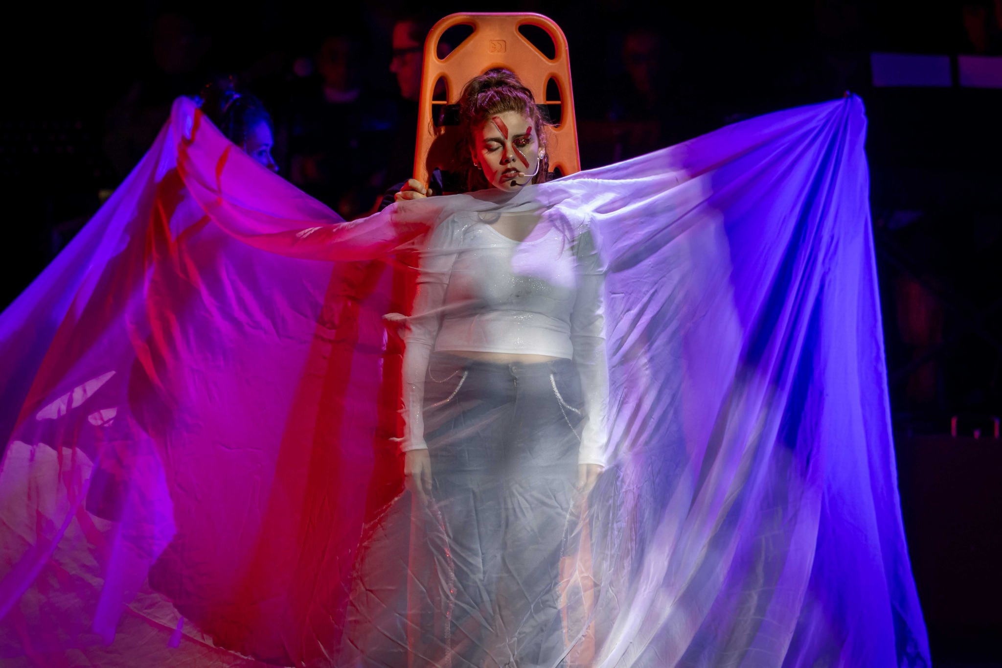 Una participante extiende una tela translúcida iluminada en azul y rosado sobre el escenario.