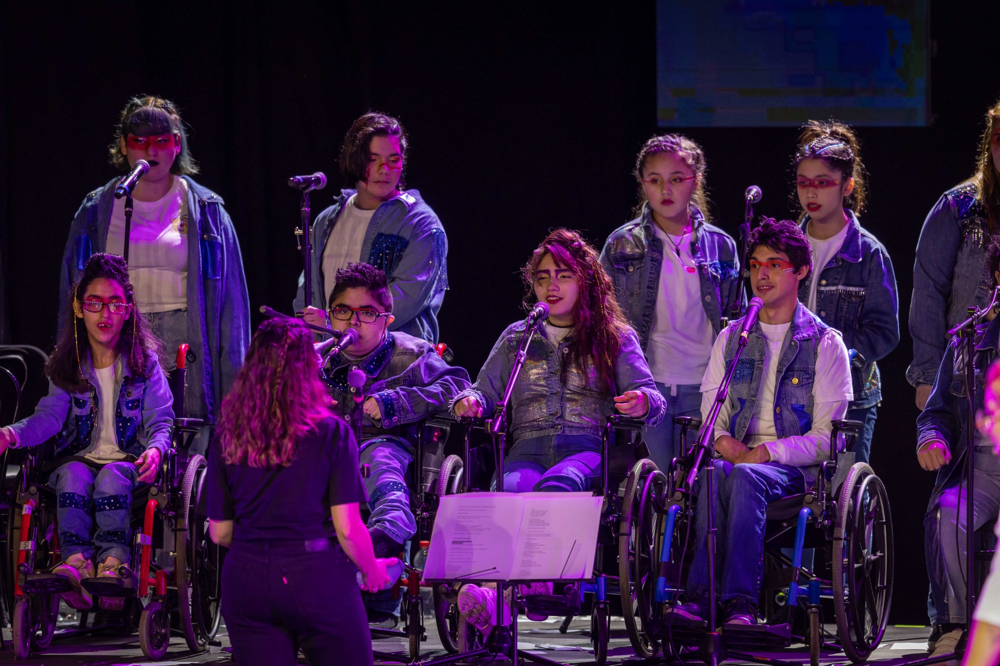 Un grupo de participantes, algunos en silla de ruedas, comparte el escenario bajo luces violetas.