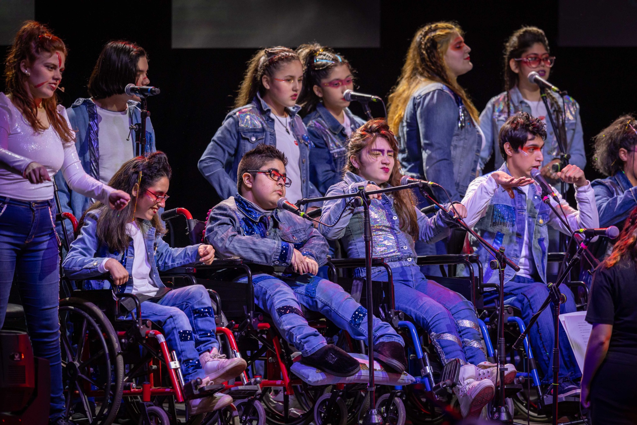 Participantes cantan ante micrófonos, algunos en silla de ruedas, bajo luces del escenario.