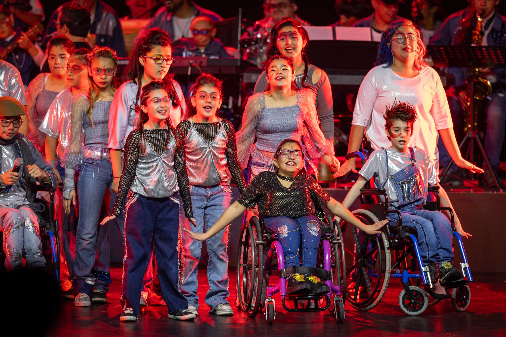 Un grupo de participantes, algunos en silla de ruedas, posa en el escenario con vestuario y luces de colores.